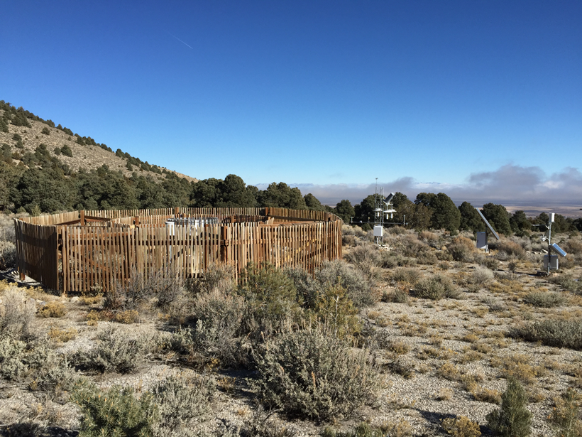 A climate reference station in the Nevada desert consisting of precipitation gauges surrounded by wooden fencing with solar-powered equipment nearby. Shrubs and brush dot the foreground and background.