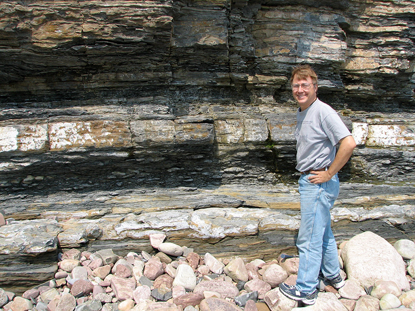 Image of Andrew Knoll standing in front of beige and gray rocks wearing jeans and a T-shirt