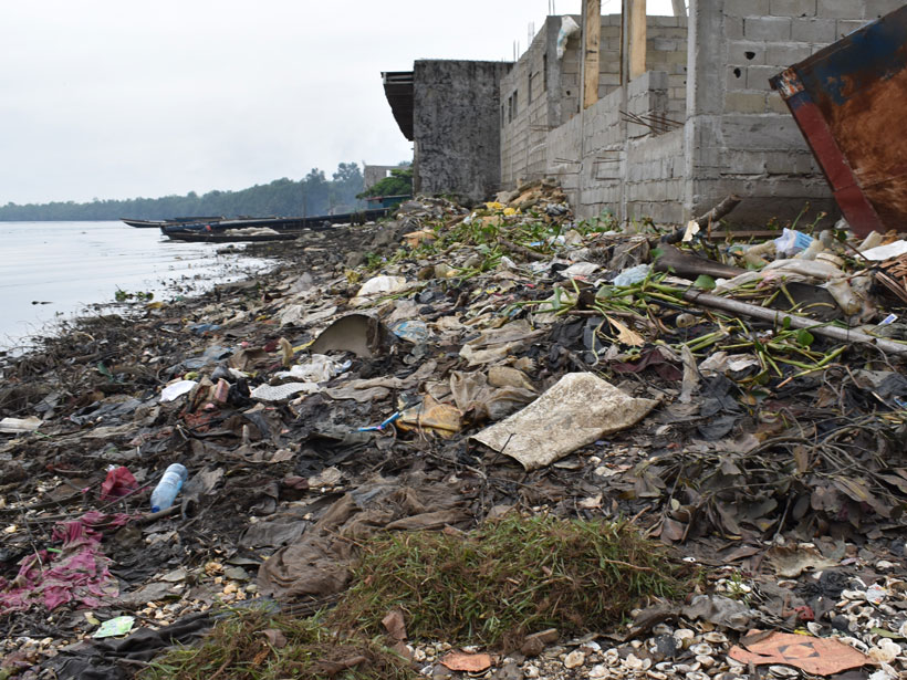 Plastic debris and other litter lines a creek shore in front of a partially constructed building