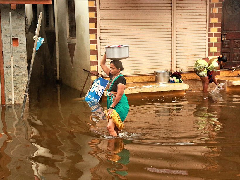 Image of a woman walking through knee-high floodwaters while balancing a large container on her head.