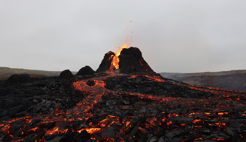 Against a gray sky, orange lava pours and pops out of Fagradalsfjall in Iceland on the second day of the eruption. In the foreground, cooling lava glows against the black basalt that’s already solidified.