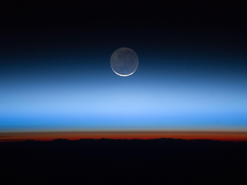 The moon appears at the top of the layers of atmosphere above the dark Earth. The orange-red glow is Earth’s troposphere, and the brown transitional layer is the tropopause.