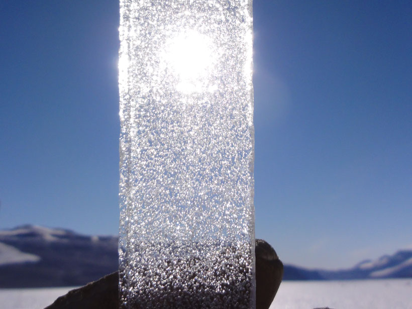 A person’s gloved hand holds part of an ice core in which air bubbles can be seen, with the Antarctic landscape in the background. The ice in the core is up to 24,000 years old.