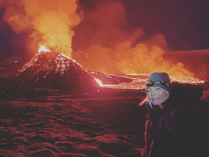 Foto de Kristin Jónsdóttir, la sismóloga de la Oficina Meteorológica de Islandia al frente, con Fagradalsfjall haciendo erupción detrás. A medida que lava naranja sale de pequeño cráter, humo anarajandizo sube al aire. El fondo de basalto negro solidificado brilla en rojo.