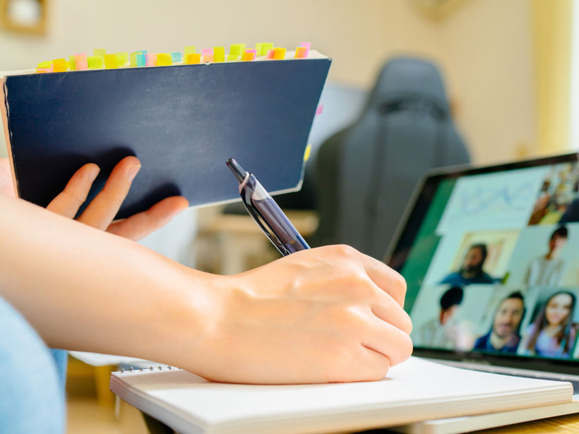Close-up view of someone’s hand as they take notes on paper during an online meeting