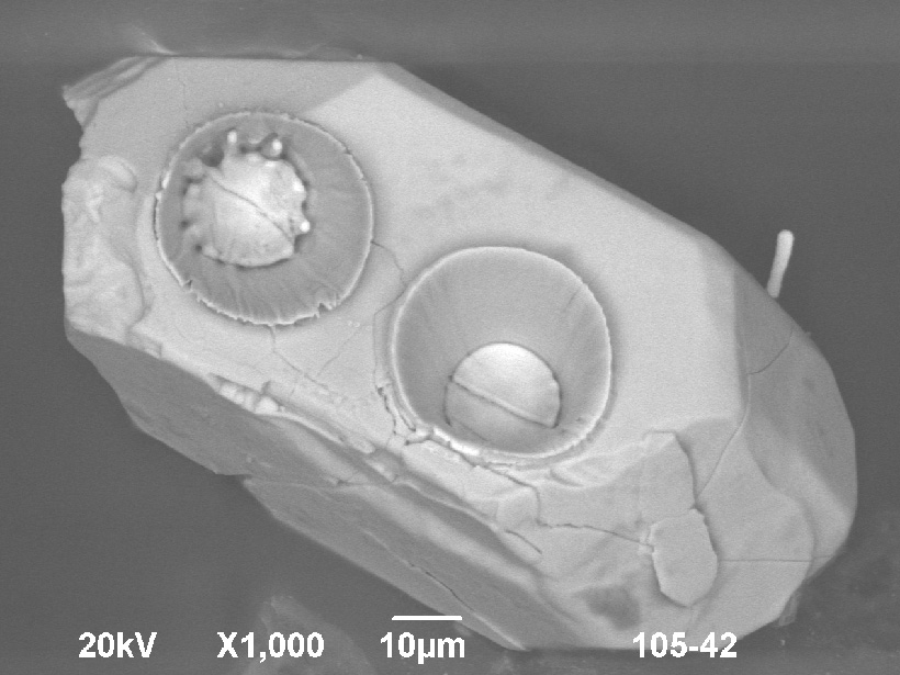 Grayscale scanning electron microscope image of an unpolished tetrahedral zircon crystal with two laser ablation pits, each between 25 and 30 micrometers in diameter