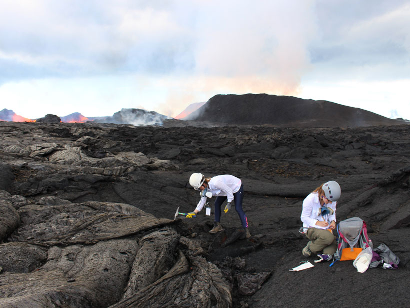 Two researchers taking samples at volcano fissure with lava in background.