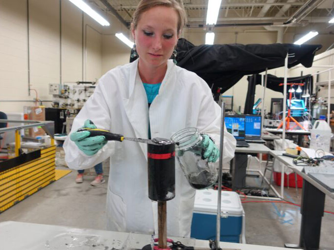 A woman in a lab coat scrapes thick material from a cylinder into a glass jar.