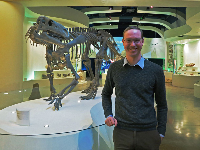 A smiling man about 30 years old stands in a brightly lit museum exhibit hall next to a meter-tall dinosaur fossil that stands on a platform.