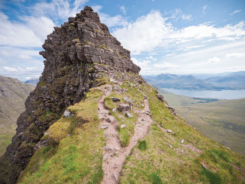 Two dirt paths in the grass diverge as they ascend a hill with a rocky peak.
