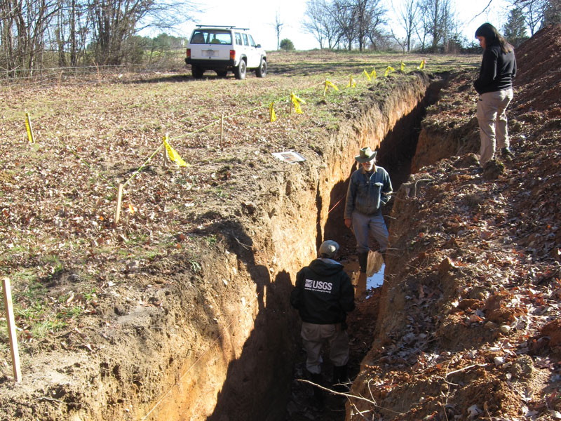 Two people stand in a deep trench in the soil while a third person stands above the trench.