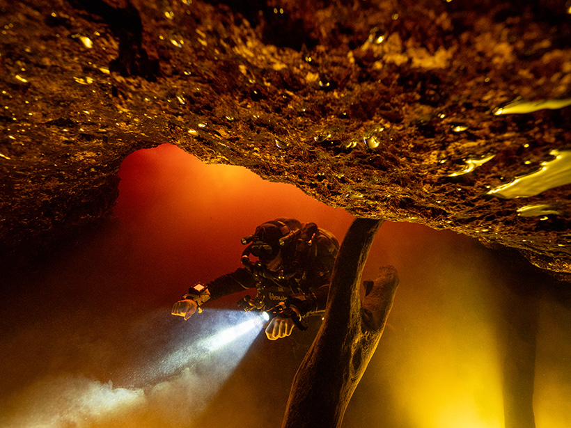 A scuba diver swims and shines a flashlight through brownish river water in a cave.
