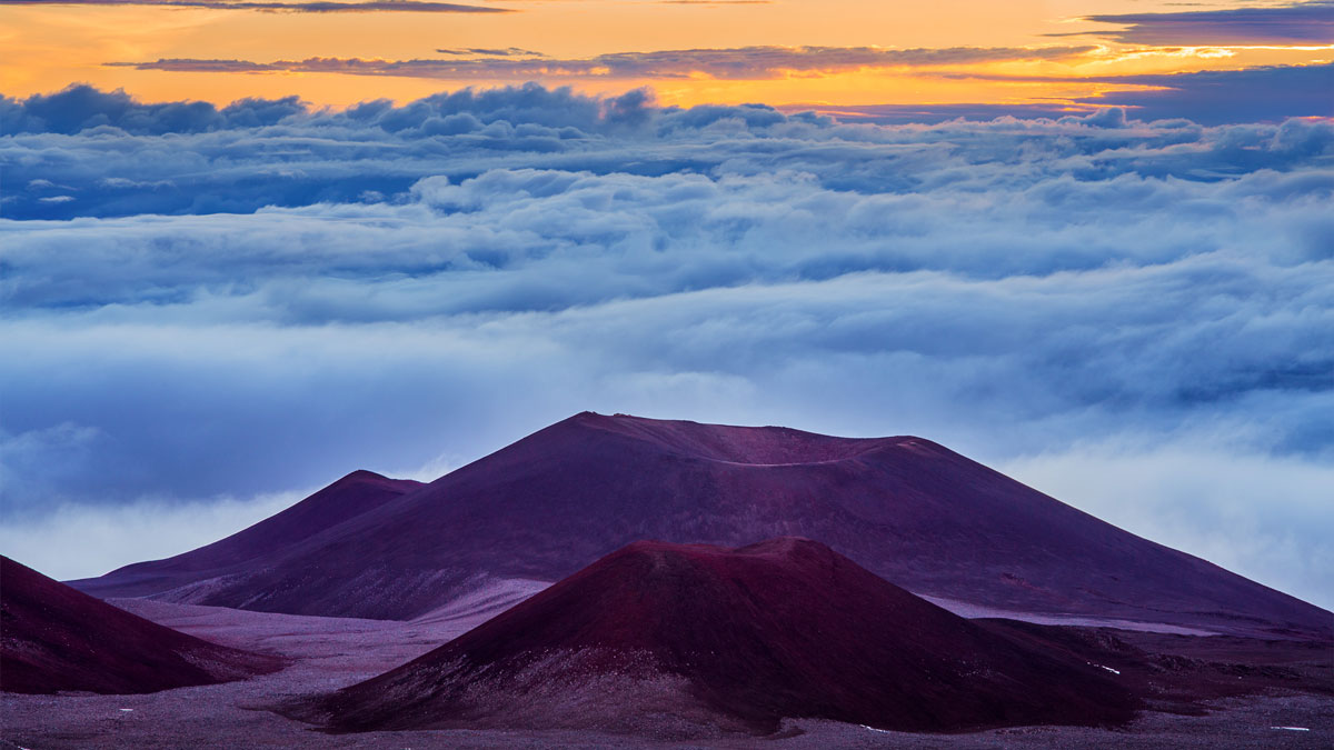 Orange sky from a sunrise fills the sky beyond a mountain summit and clouds.