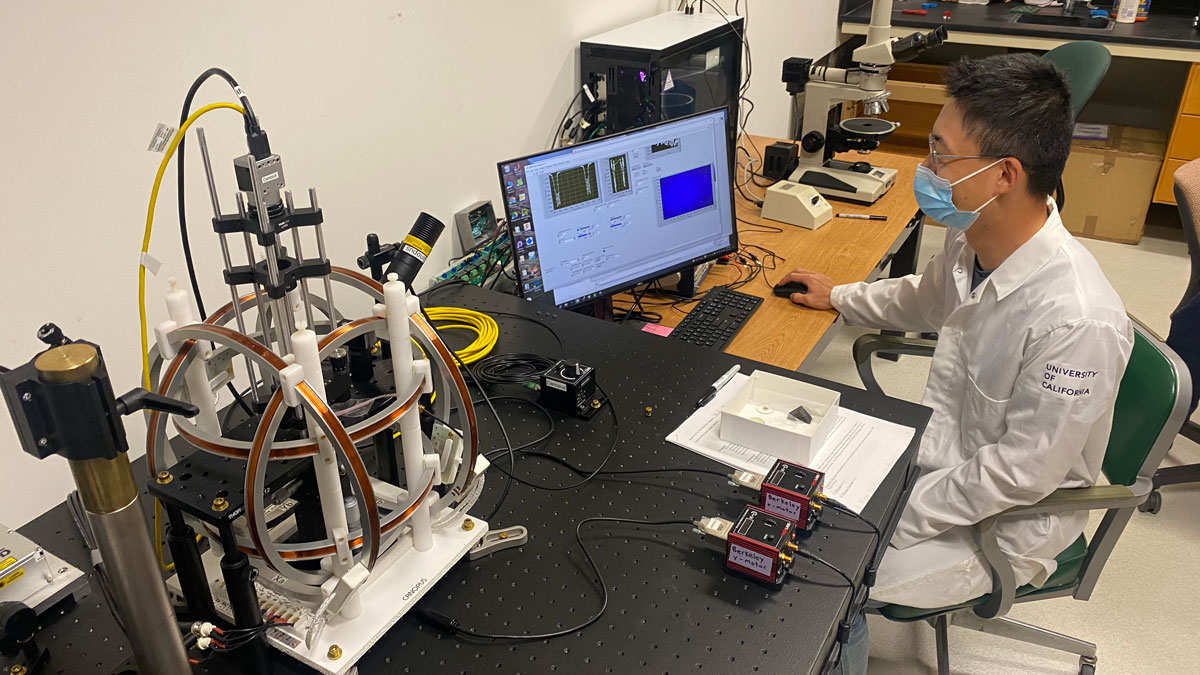 Wearing a white lab coat, Yiming Zhang, a doctoral student at the University of California, Berkeley, sits in front of a computer screen, examining data, with a mouse in his right hand. To his left, a gray microscope with four copper-colored rings encircling the stage perches on a black table.