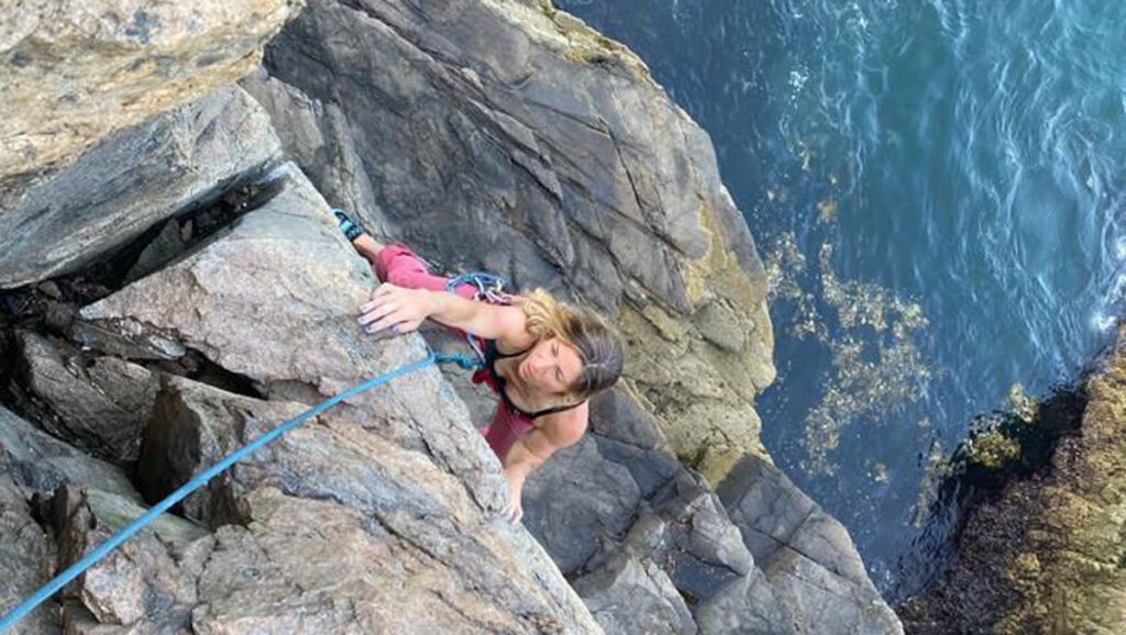 A woman rock climber reaches over a granite ledge with the ocean below in Acadia National Park.