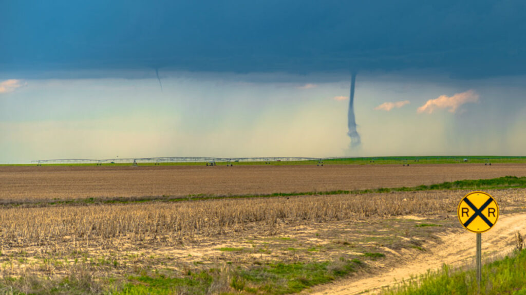 Looking across Midwestern cropland, the viewer sees a tornado extending down from thick, gray-blue clouds to meet the horizon. To the tornado’s left, a funnel cloud companion looks like a thin finger pointing toward Earth from the bottom of the clouds.