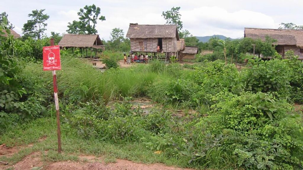 In the foreground are tall grasses and green leafy bushes that cover hidden mines. A red sign with a skull and crossbones reads “Danger! Mines!” in both English and Khmer. Three wooden houses on stilts sit in the background of the image, and a group of villagers is gathered around the base of the central house. More houses extend into the distance, and a mountain range is visible on the horizon.