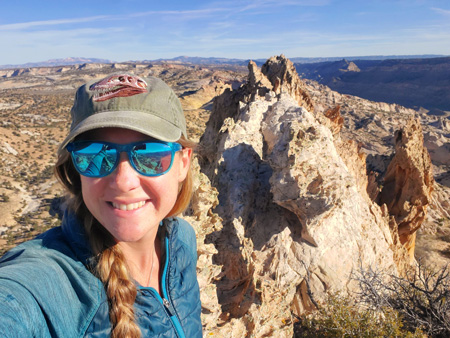 A woman with a tyrannosaur skull on her hat smiles at the camera in front of a jagged landscape of spires, cliffs and canyons.