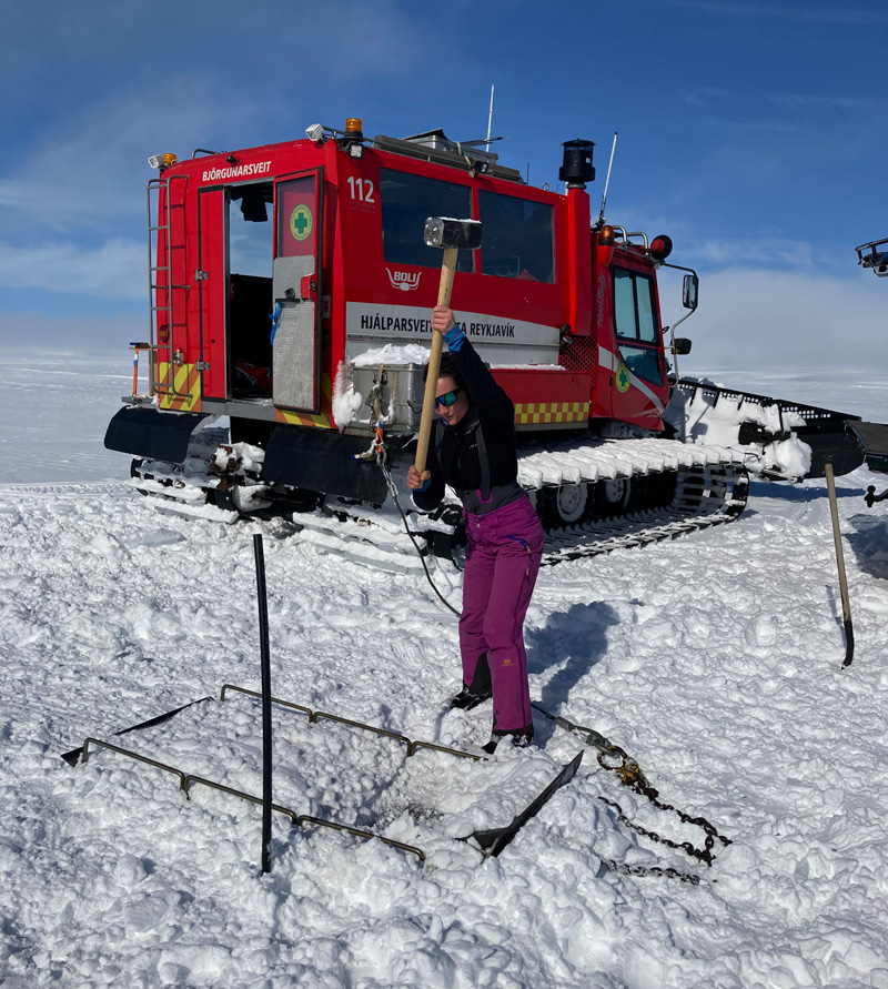 A researcher pounds on a metal frame in the snow with a sledgehammer in front of a snowcat.