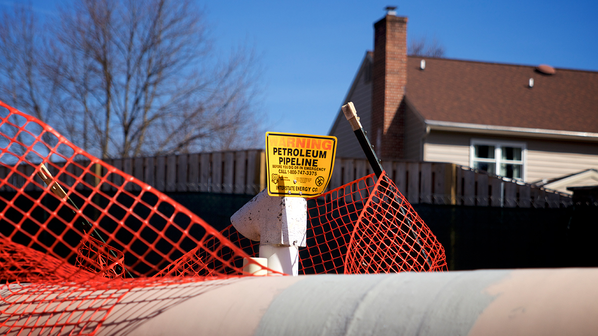 A gray and tan pipeline crosses horizontally along the bottom of the image with a bright orange plastic construction fence draped over it. A yellow sign reading “Warning: Petroleum Pipeline” sits in the center of the image. In the background are a tall wooden fence, the top of a two-story house, bare trees, and a clear blue sky.