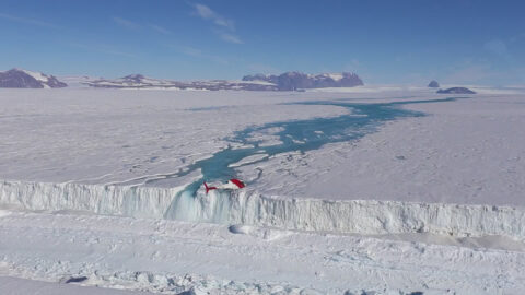 A helicopter hovers in the foreground as meltwater pours from a waterfall over the edge of an ice shelf.