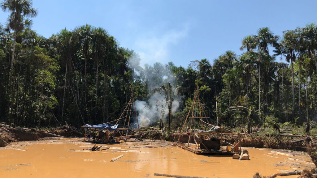 In the foreground sits a yellow-beige pond, with tree trunks scattered about its surface. Two dredges that appear to be made of wood float by the banks, smoke rising from between them. In the background, the green rain forest towers, the blue sky smudged with white smoke.