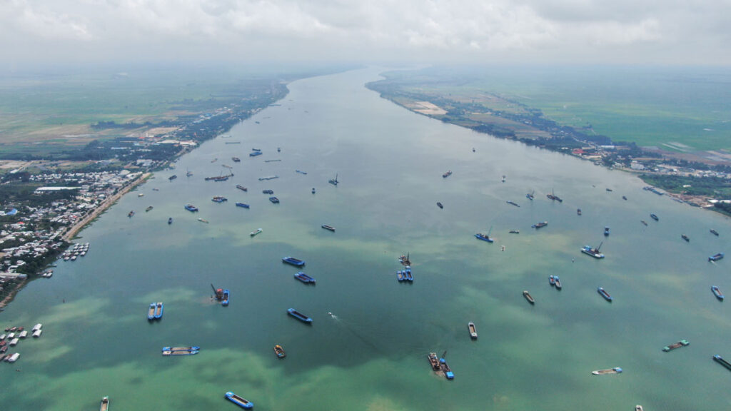 Under a cloudy sky, numerous blue-tipped sand mining ships dot the green-blue waters of the Mekong River.