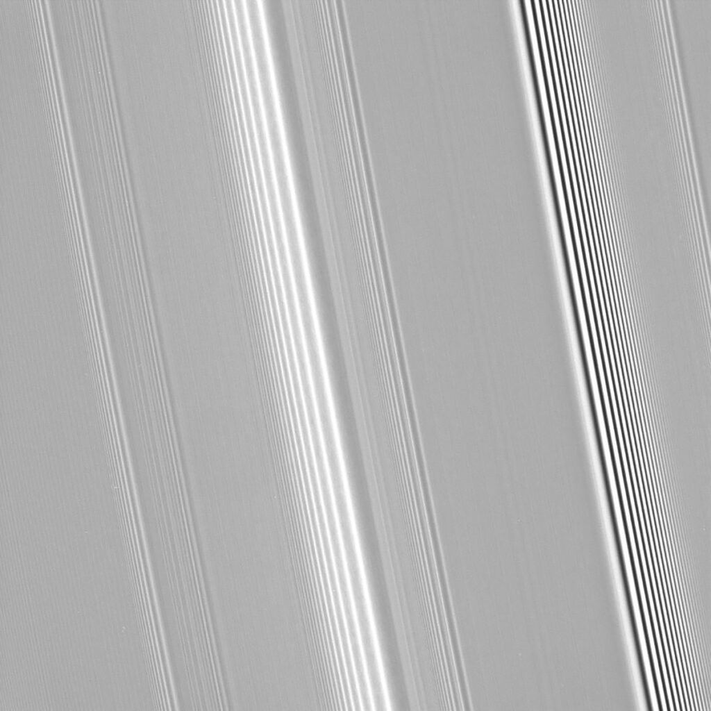Saturn’s rings streak across the image diagonally from top left to bottom-right in varying shades of gray. Lighter stripes represent more loosely packed rings, and darker stripes are more densely packed rings. At the center is a bright wave pattern, and toward the right is a darker wave pattern.