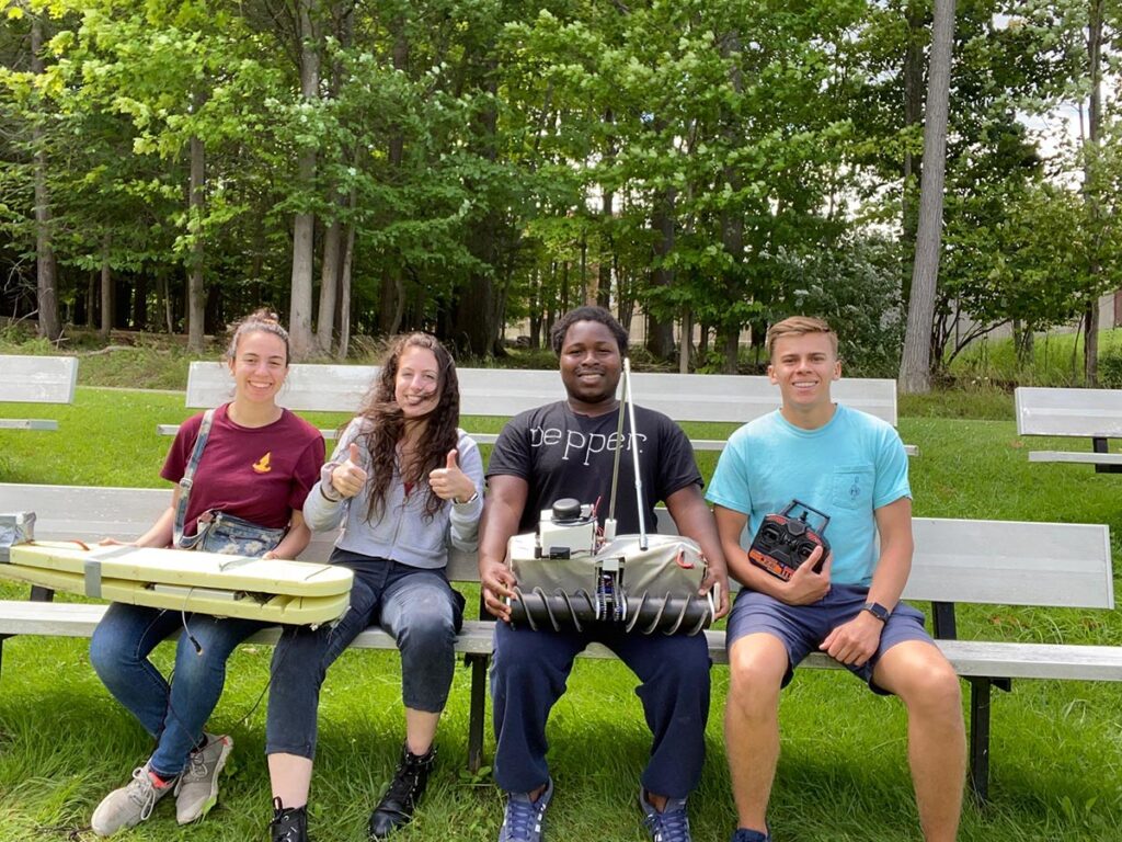Four smiling people sit on a bench on a sunny day in a grassy park with trees in the background. The two white women on the left are holding a surfboard-shaped sensor array in their laps. Second from the right sits a black man holding a small robot with a tall antenna. On the left sits a white man holding a remote controller that steers the robot. All of the people are wearing casual clothing.