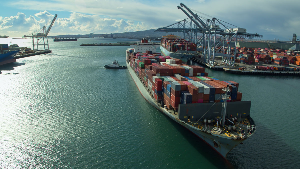 A large, fully loaded container ship arrives at a busy port in California.