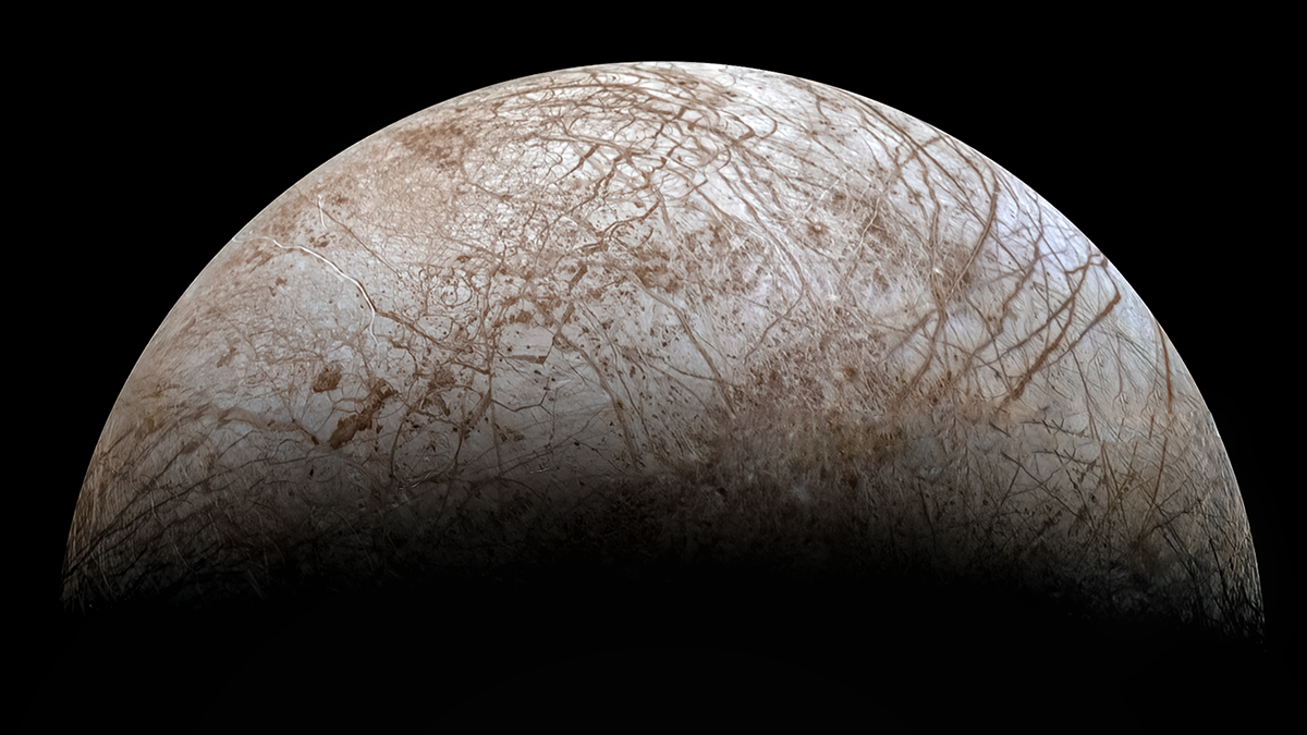 A hemisphere of the moon Europa sits on a black background, with the curve of the horizon toward the top of the image. The moon’s surface is a light gray-brown and is crisscrossed with dark brown streaks and splotches.