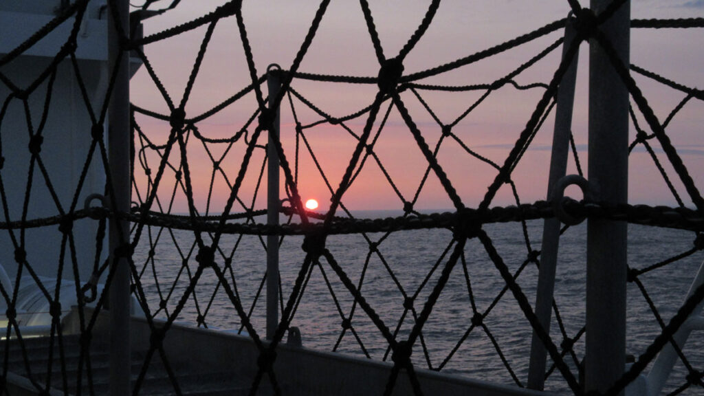 A sunset over the ocean is seen through rope netting on a ship