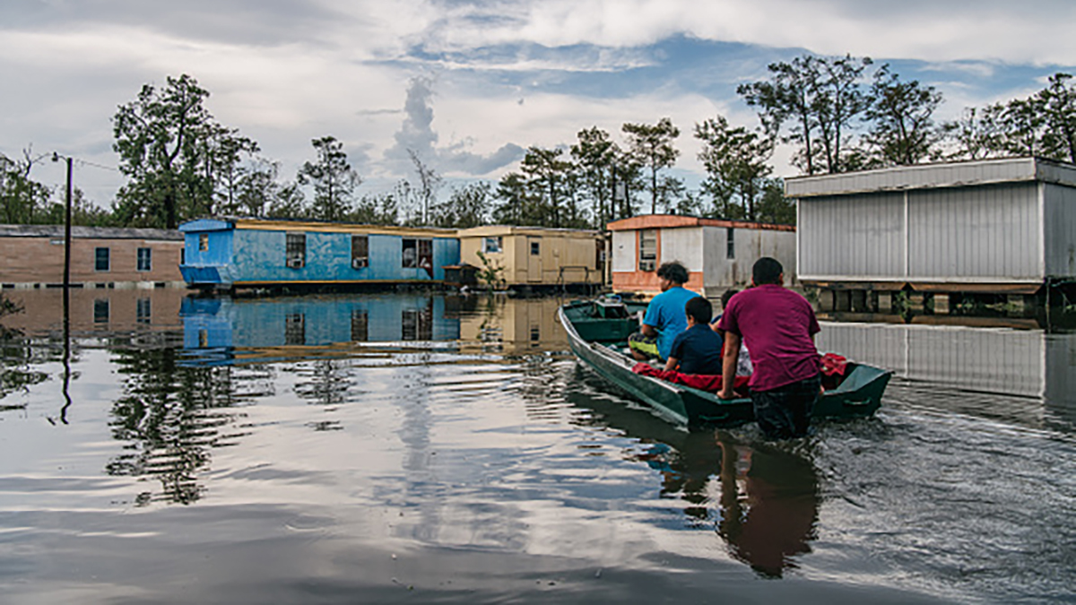 Four Black people sit in a small motorized boat as it moves up a flooded street toward prefabricated and mobile homes. The boat is near the bottom right of the image and is pointed toward center-left. All four people face away from the viewer. The sky is blue but mostly clouded over.