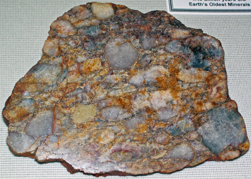 A polished flat cut of rock sits on a white fabric background with the label “Earth’s Oldest Minerals” in the upper right. The cut spans the width and height of the image and has smooth but irregular edges. The rock is composed of smaller orange, brown, and blue rock fragments of a range of sizes that are tightly packed together.