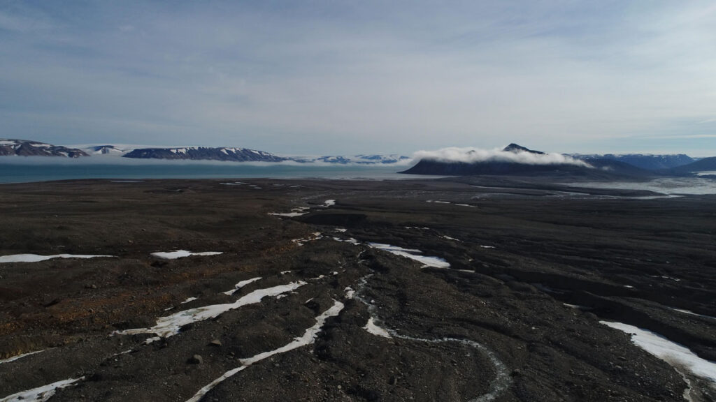 Brown, barren, relatively flat land stretches into the distance, dotted with occasional patches of white snow. The dark blue Arctic Ocean laps the shore. A thin sliver of sky is gray and cloudy.