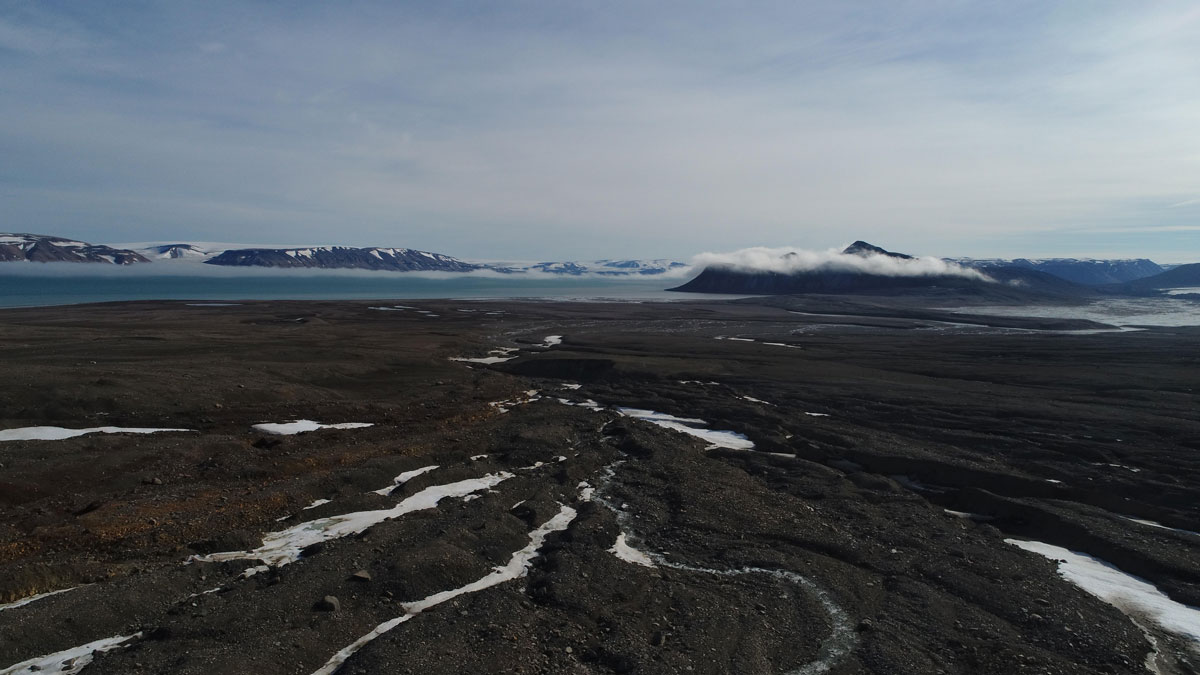 Brown, barren, relatively flat land stretches into the distance, dotted with occasional patches of white snow. The dark blue Arctic Ocean laps the shore. A thin sliver of sky is gray and cloudy.