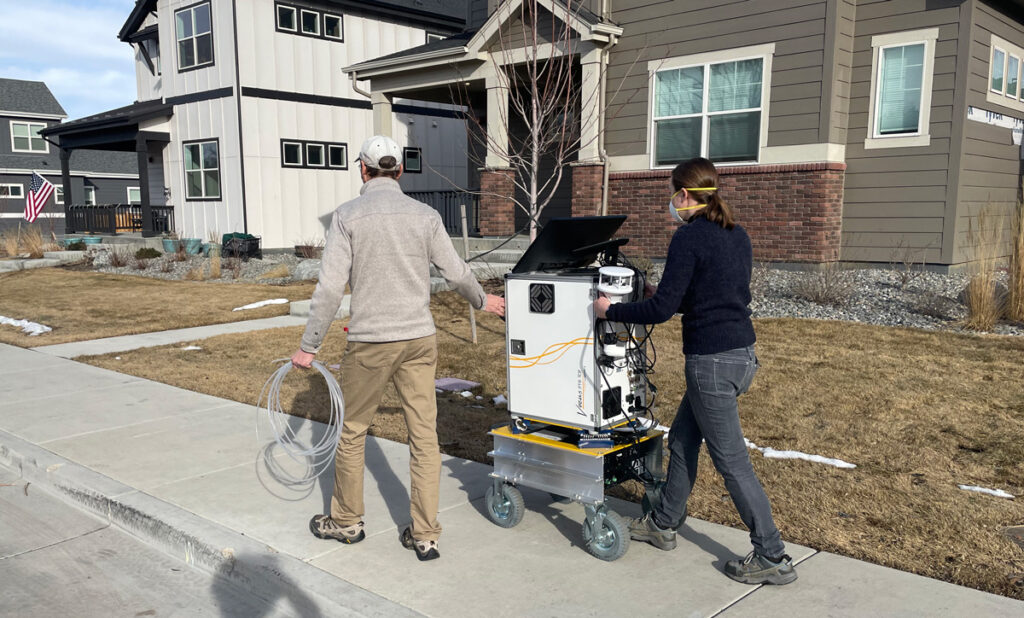 Two people guide a trolley carrying a white instrument with black wires and buttons on its back along a sidewalk past houses.