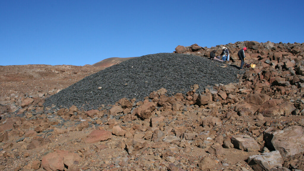 Under a clear blue sky, a field strewn with brown volcanic rocks contains a pile of black basalt that rises like a giant anthill, with several people on the right edge for scale.