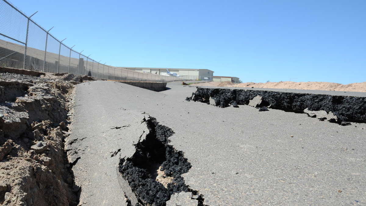 Camino dañado por un terremoto en Calexico, California, 2010. El camino muestra grietas en el pavimento. Al fondo se observa un edificio de una planta cercado y el cielo azul.