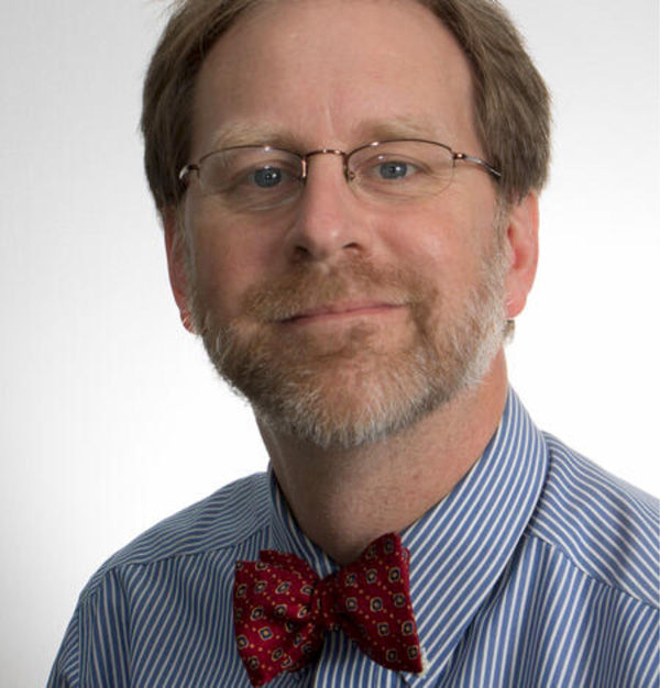 A headshot of a middle-aged white man from the shoulders up in front of a white background. The man, David Applegate, wears a blue stripped shirt, a red bow tie, and thin-framed glasses. Applegate has light brown hair, a short brown-gray beard, and light colored eyes.