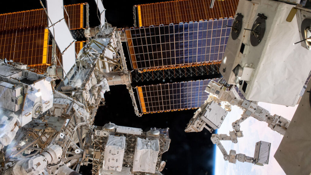 In the foreground, the GEDI instrument appears as a large white box. In the background, an astronaut climbs along the space station’s external scaffolding.