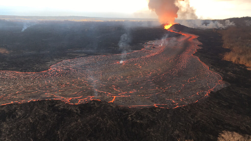 A river of molten lava flows across a steaming black basalt landscape. The river flows from a volcanic rift near the top right of the image toward the bottom left. The fissure is filled with brighter and hotter lava and steam and gas billows up from it. The sky on the horizon is a hazy blue-gray.
