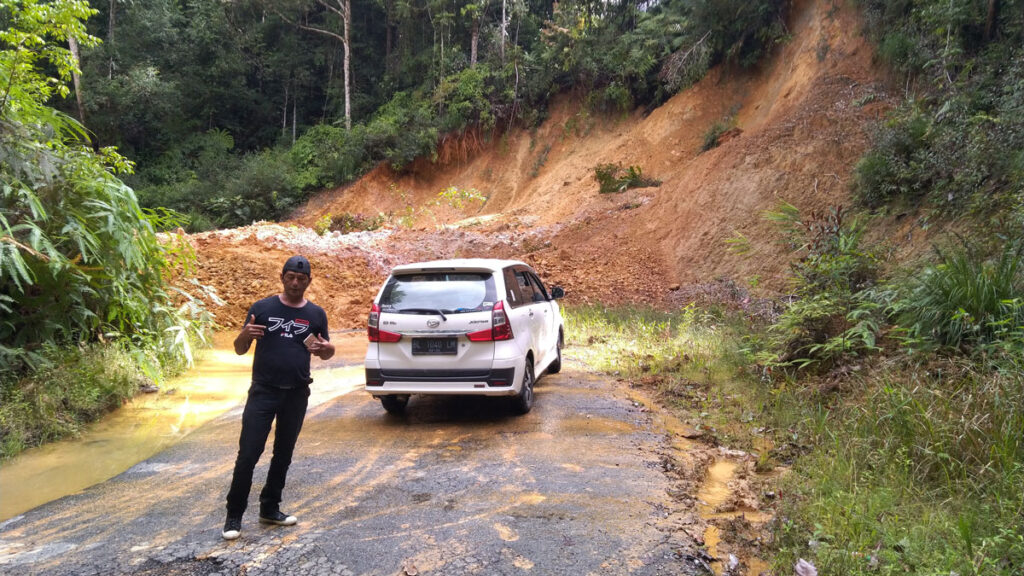 A man stands in front of a car on a road blocked by a large pile of soil from a landslide.