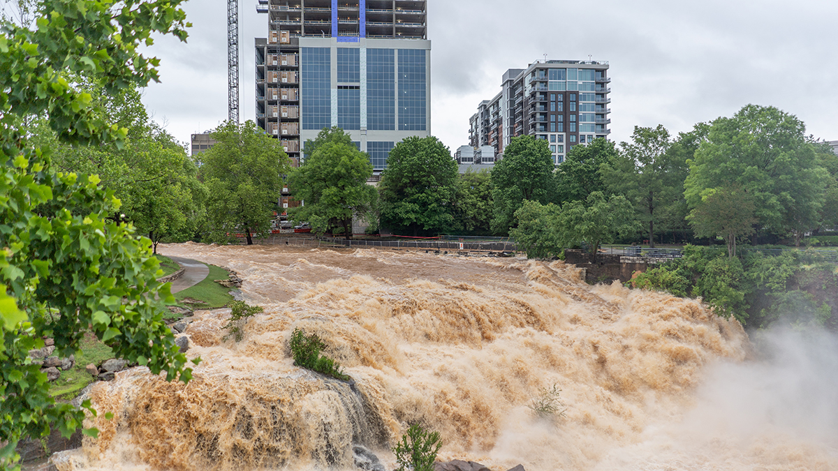 Brown river water rushes through a downtown area with industrial and residential buildings in the background.