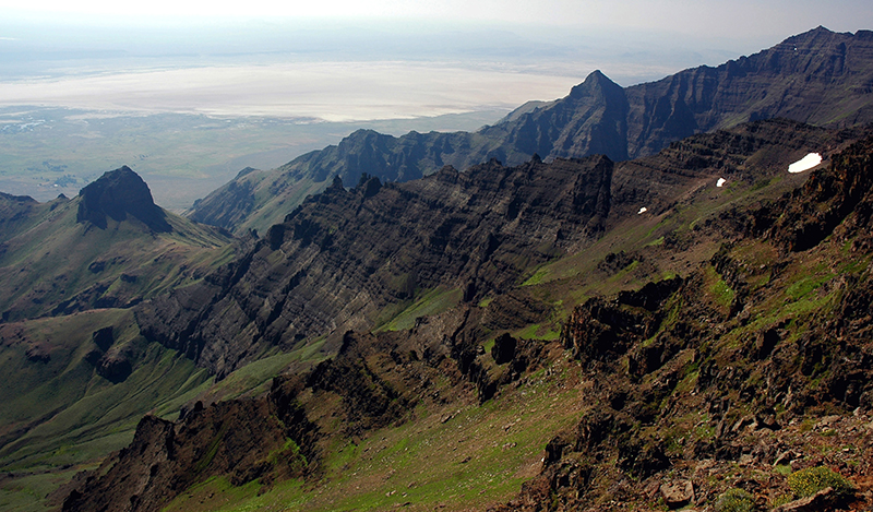 Stratified basalt mountain ridges partially covered with green vegetation are visible from the summit of Steens Mountain. The lower-right diagonal half of the image shows the mountainside ridges and the upper-left diagonal half shows further basalt formations in the distance.