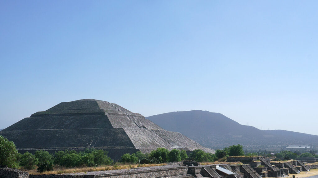 La pirámide del Sol en Teotihuacan al frente con un cerro y el cielo despejado detrás.