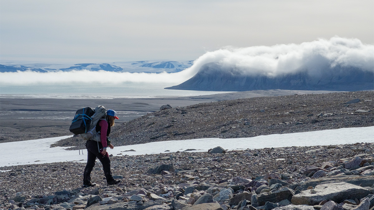 The article author, carrying a pack and equipment on her back, hikes across a rocky expanse with mountains and low-lying clouds in the background.