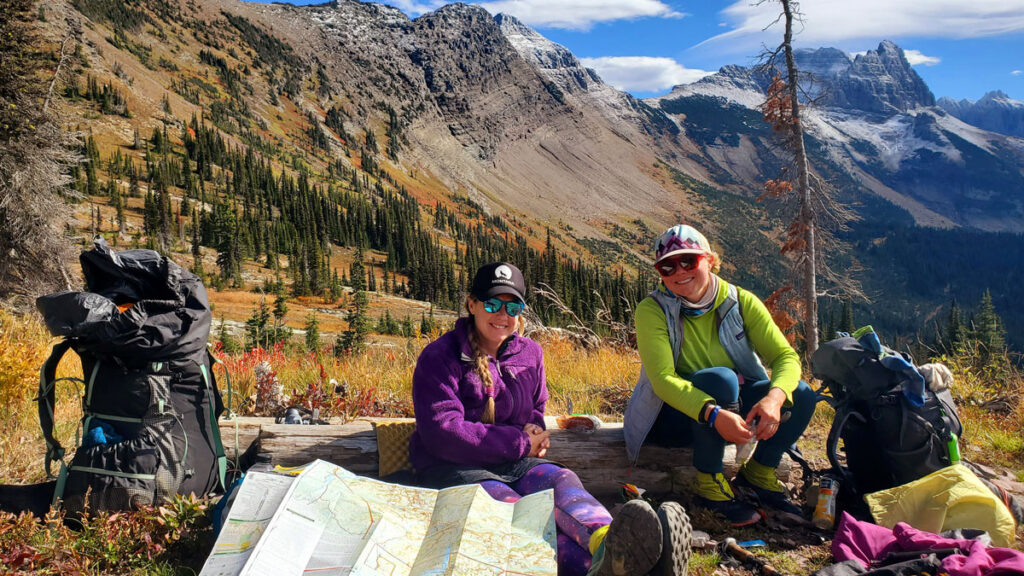 Two women sit on a log next to their backpacks and a map, with a snowy ridge of mountain peaks in the background.
