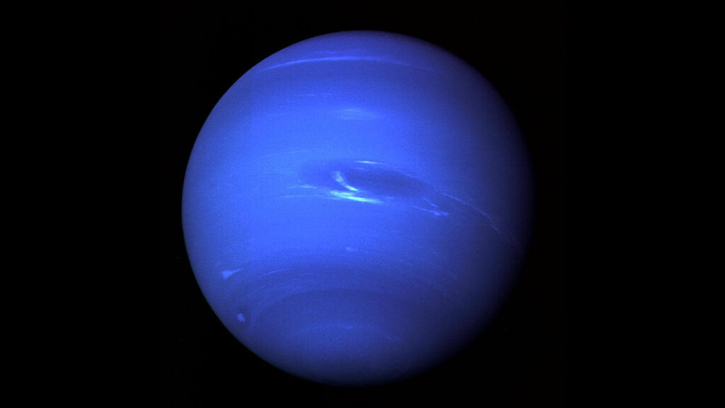 The dark blue orb of Neptune is viewed by Voyager 2 at an upward angle from the south pole. A dark navy storm spot, the Great Dark Spot, is just to the right of the center of the planet, and white high-altitude clouds are scattered around the planet.