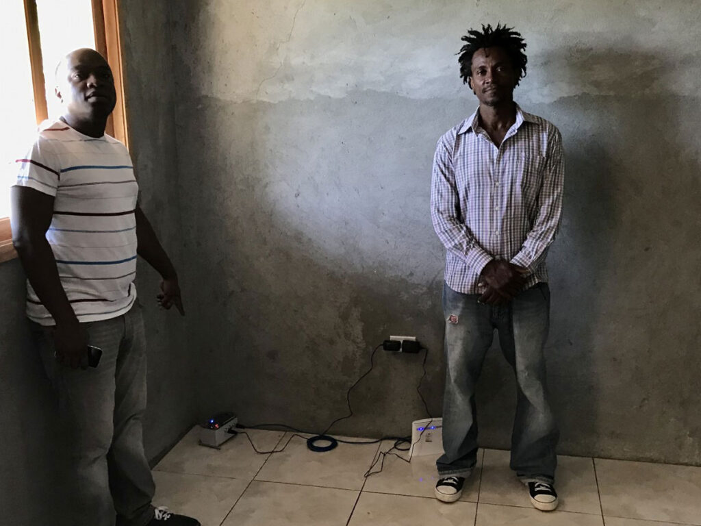 Two men stand in a room. The man on the left points to a Raspberry Shake seismometer, the small box on the floor.